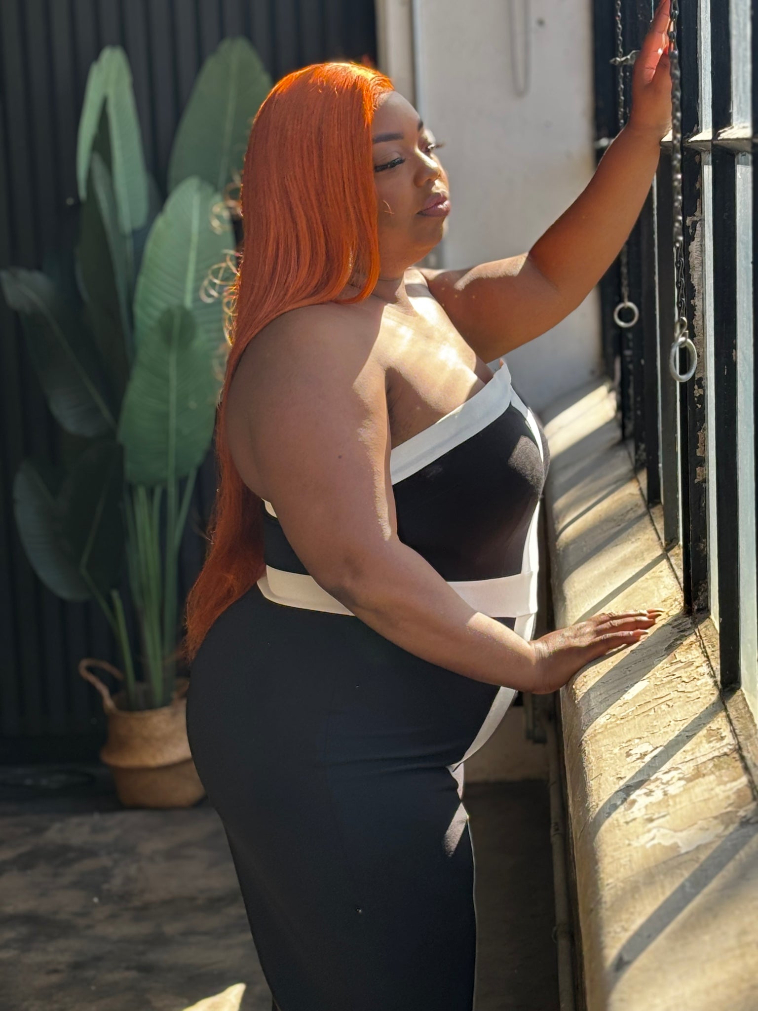 Plus size woman wearing a strapless black and white colorblock jumpsuit with wide legs, stretchy fabric, and a side zipper, standing confidently in a modern maroon and gold-accented room